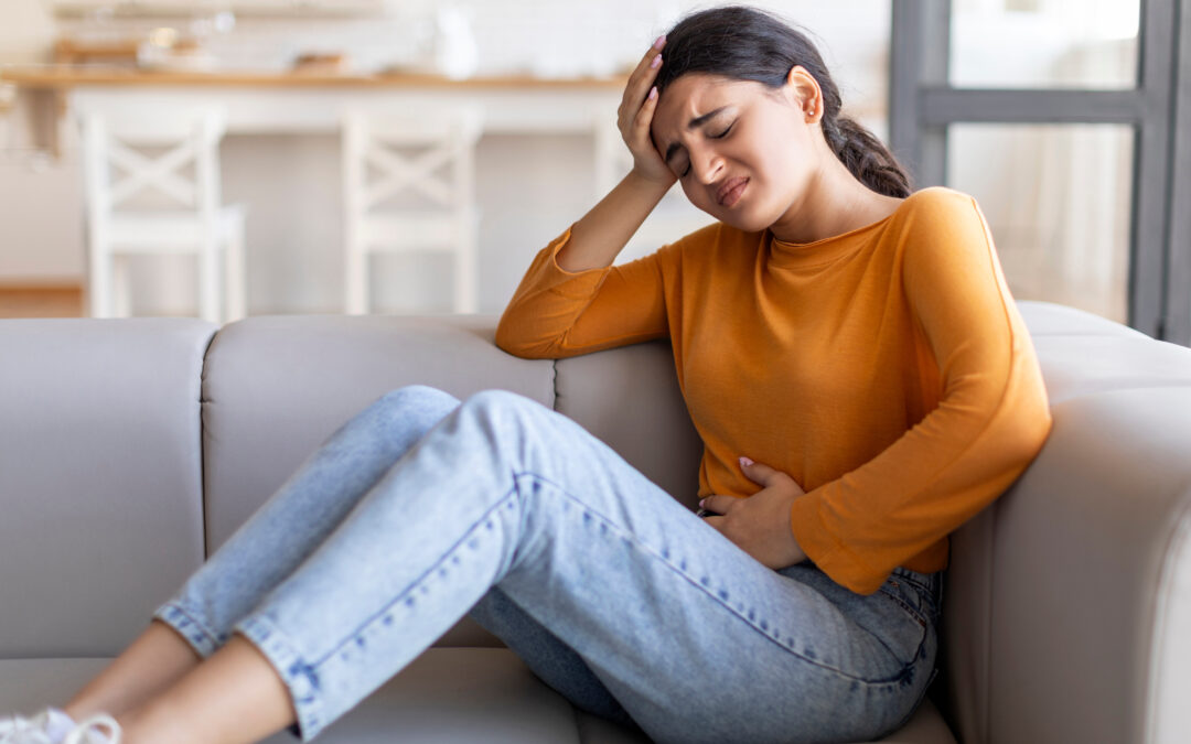 Woman in orange shirt and jeans sitting on couch holding her stomach and head in pain, showing symptoms of abdominal distress possibly related to a norovirus infection.